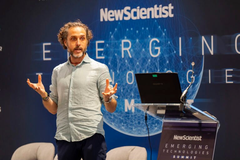 A person with curly hair speaks enthusiastically on stage at the New Scientist Emerging Technologies Summit. A laptop is on a transparent podium in front of them, and the event's logo is displayed on a large screen behind