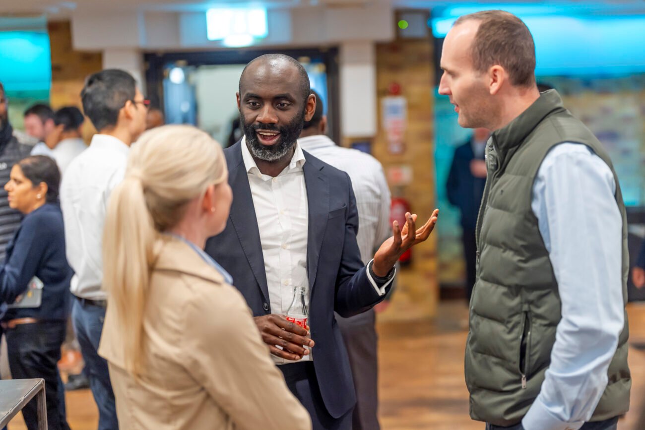A group of people engaging in conversation at a social event. A man in a suit gestures animatedly while holding a drink, accompanied by a woman in a beige coat and a man in a vest with folded arms. Other attendees are visible in the background. James Gifford-Mead Photography - Event Photographer London