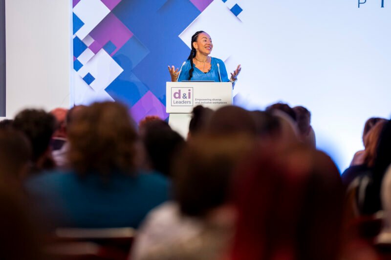 A woman stands at a podium, smiling and gesturing with her hands during a presentation. The audience is seated in the foreground. The background features a geometric design. James Gifford-Mead Photography - Event Photographer London