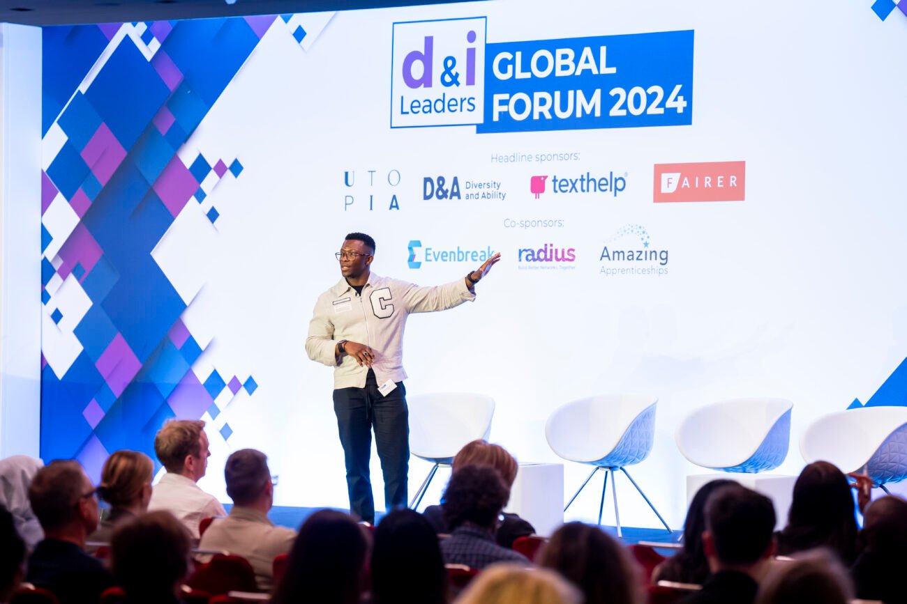 A speaker stands on stage at the d&i Leaders Global Forum 2024. They gesture towards a projected screen displaying event and sponsor logos. The audience is seated and listening attentively. James Gifford-Mead Photography - Event Photographer London