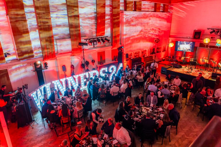 A large event with people seated at tables adorned with drinks and flowers. A stage with instruments is seen in the background. The room is illuminated with red and white lighting, showcasing large lit letters spelling "WINNERS"