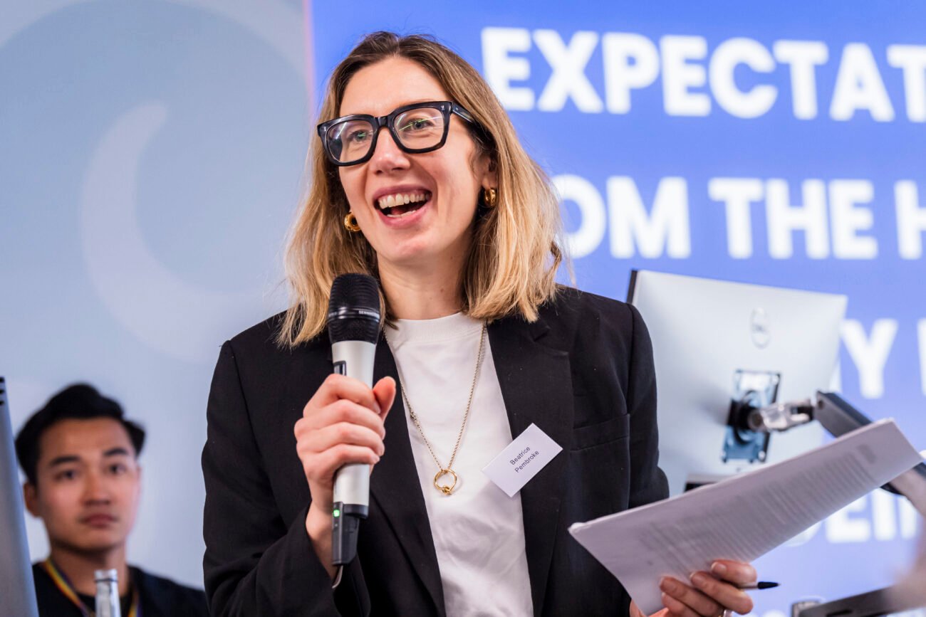 A person with glasses and a black blazer holds a microphone and papers, smiling while speaking. In the background, a person listens, and part of a blue wall with partially visible text is shown. James Gifford-Mead Photography - Event Photographer London