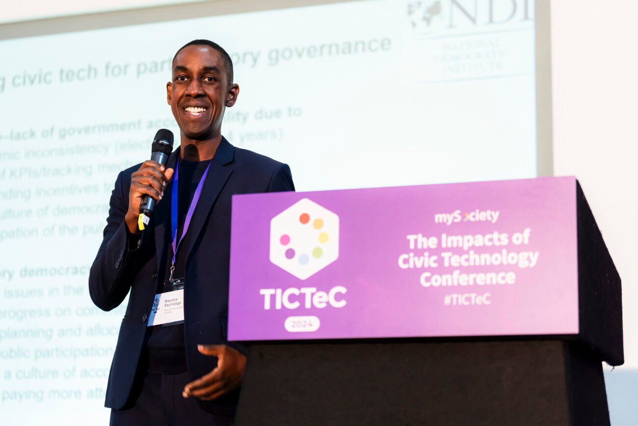 A speaker smiles while holding a microphone at the TICTeC 2024 conference. He's standing next to a podium with a slide presentation in the background. James Gifford-Mead Photography - Event Photographer London