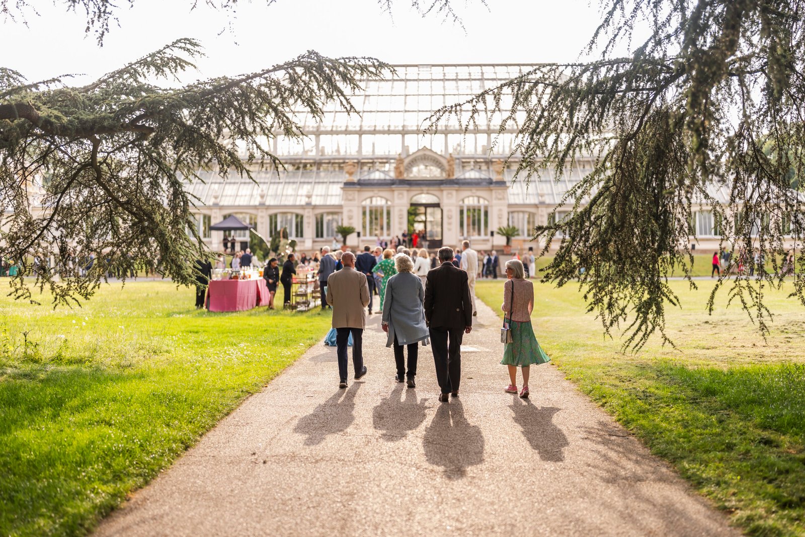 A group of people walks along a path leading to a large glass greenhouse on a sunny day. The greenhouse is surrounded by trees, and a pink table is visible on the lawn. The scene suggests a social gathering. James Gifford-Mead Photography - Event Photographer London