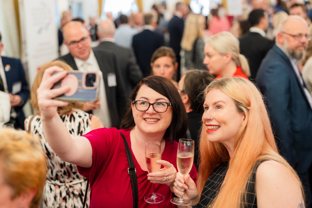 Two people in a crowd take a selfie while holding champagne glasses. They are smiling, and the background is filled with people engaged in conversation, suggesting a lively social event or celebration. James Gifford-Mead Photography - Event Photographer London