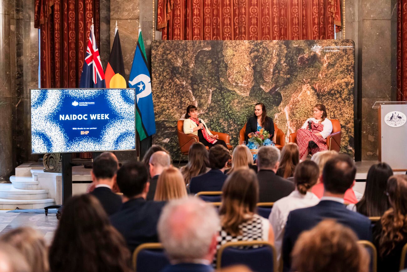 Three people sit on stage in discussion chairs, with a screen displaying "NAIDOC Week" beside them. Flags and a large map serve as the backdrop. Audience members are seated in the foreground. James Gifford-Mead Photography - Event Photographer London