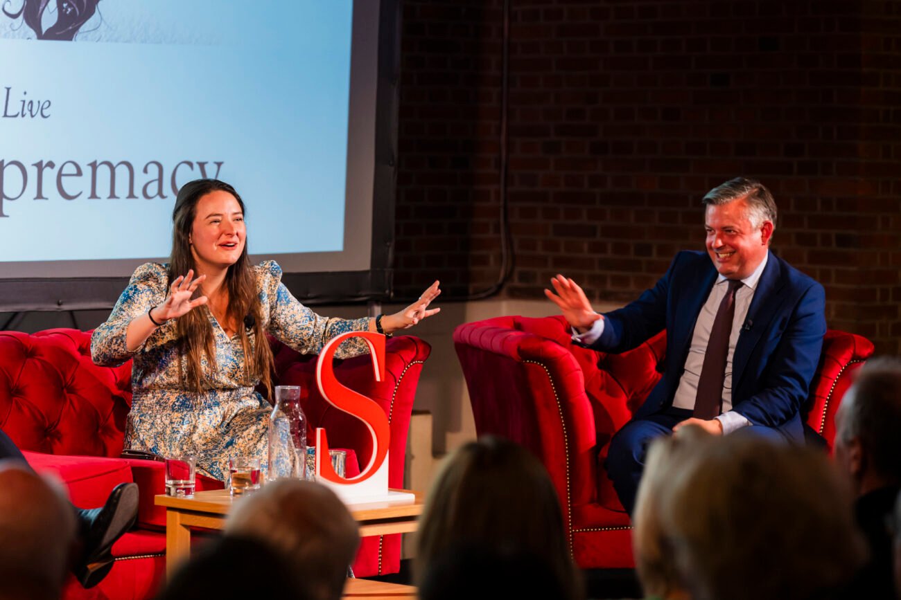 Two people sit on red couches during a discussion on stage. The woman gestures with her hands, wearing a patterned dress. The man in a suit smiles. A slide is projected behind them with partial text visible. Audience members are in the foreground. James Gifford-Mead Photography - Event Photographer London