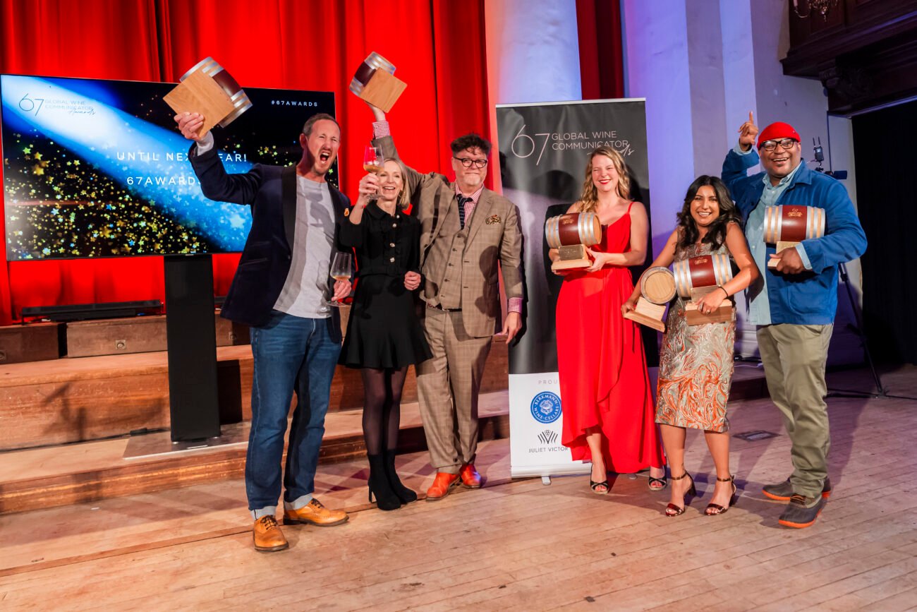 A group of six people stands on a stage, each holding a small barrel-shaped award. They are smiling and celebrating. A large screen and a banner in the background indicate a formal event with bright red curtains behind them. James Gifford-Mead Photography - Event Photographer London