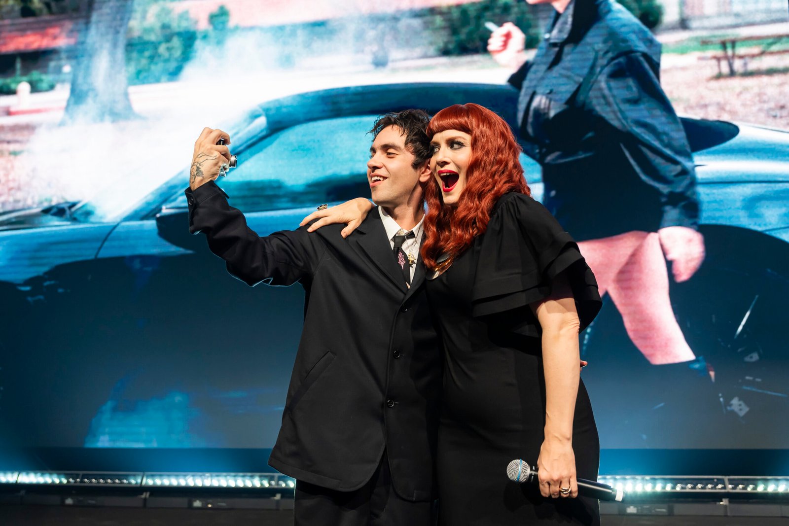 A man and a woman with red hair are taking a selfie on stage, smiling. They are dressed in formal black attire, standing in front of a large screen displaying a scene with a car and a person in the background