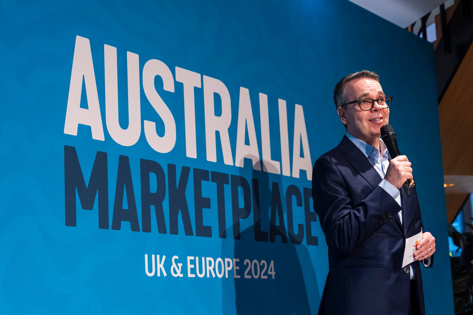 A person in a suit speaks into a microphone next to a large blue sign that reads "AUSTRALIA MARKETPLACE UK & EUROPE 2024