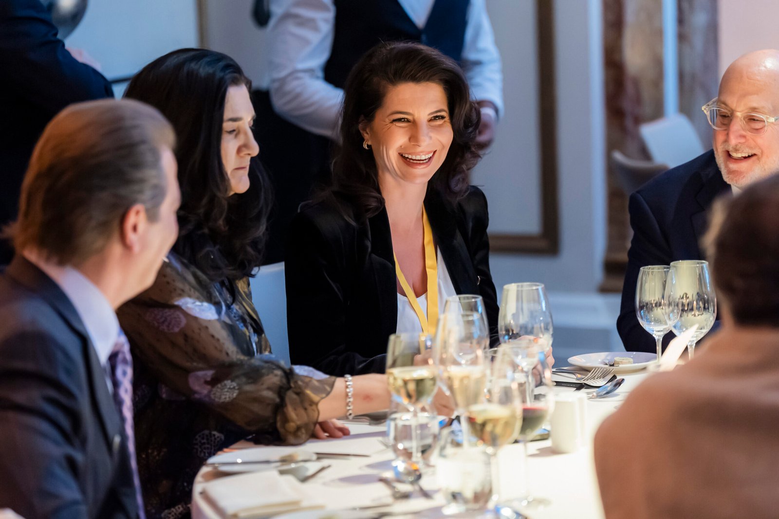 A group of people are sitting around a table, elegantly dressed, engaging in conversation and laughter. The table is set with wine glasses and plates, suggesting a formal dinner or event atmosphere