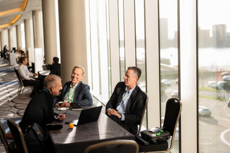 Three men in business attire sit at a table with laptops and papers in a sunlit conference hallway. Large windows reveal a cityscape. The setting appears professional, with others in the background having discussions