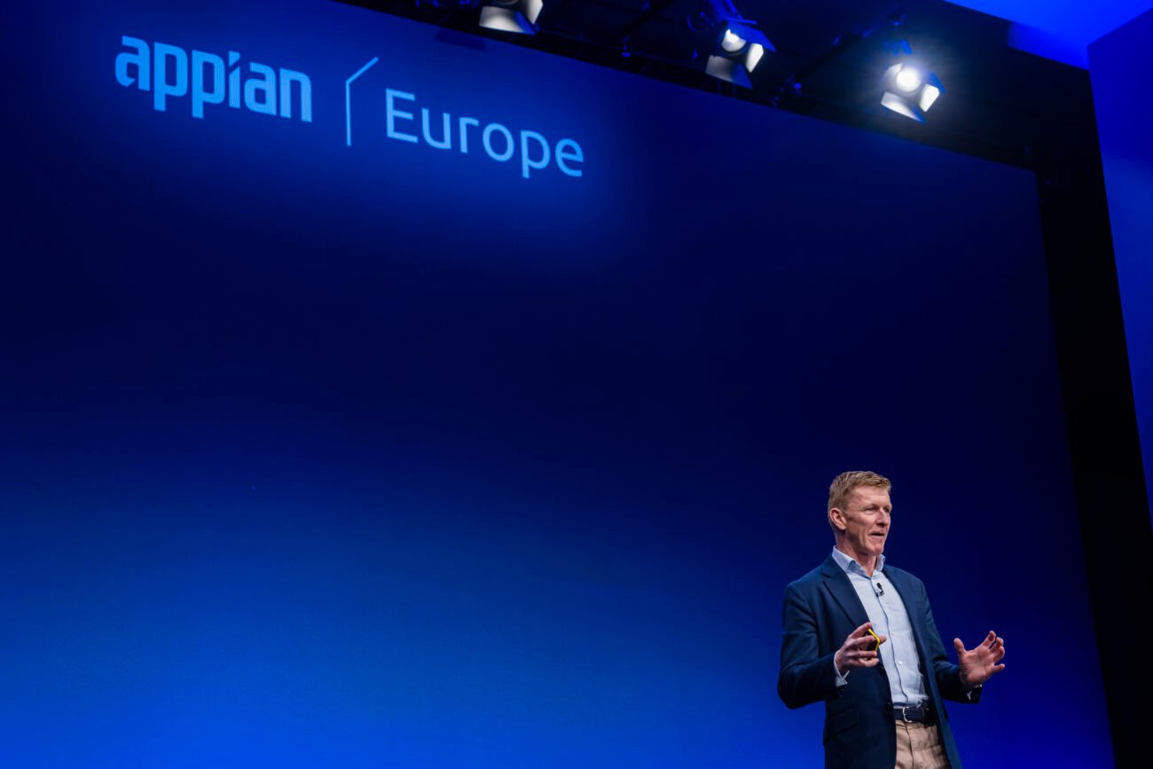 A speaker stands on stage, gesturing with his hands, under bright spotlights. The backdrop features the words "appian" and "Europe" on a blue gradient screen. The setting appears to be a conference or presentation. James Gifford-Mead Photography - Event Photographer London