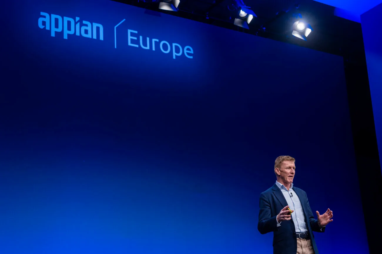 A speaker stands on stage, gesturing with his hands, under bright spotlights. The backdrop features the words "appian" and "Europe" on a blue gradient screen. The setting appears to be a conference or presentation