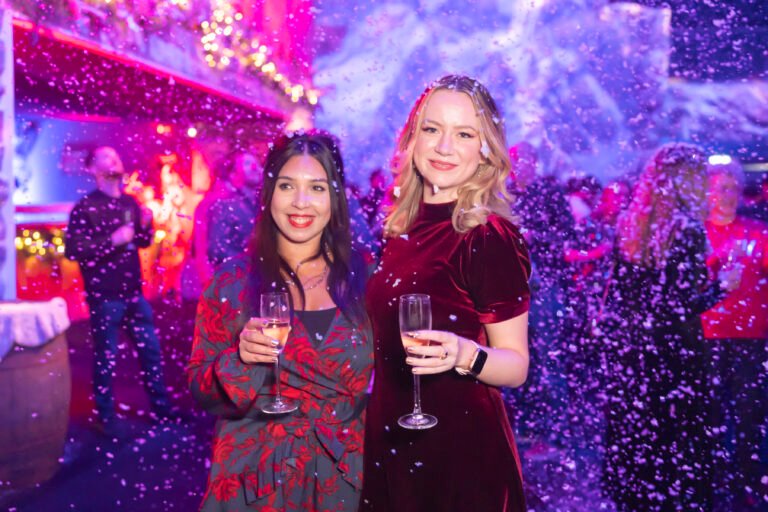 Two women in festive attire hold champagne glasses at a holiday-themed event. Snowflakes fall around them. The background is filled with people, lights, and holiday decorations, creating a joyful atmosphere