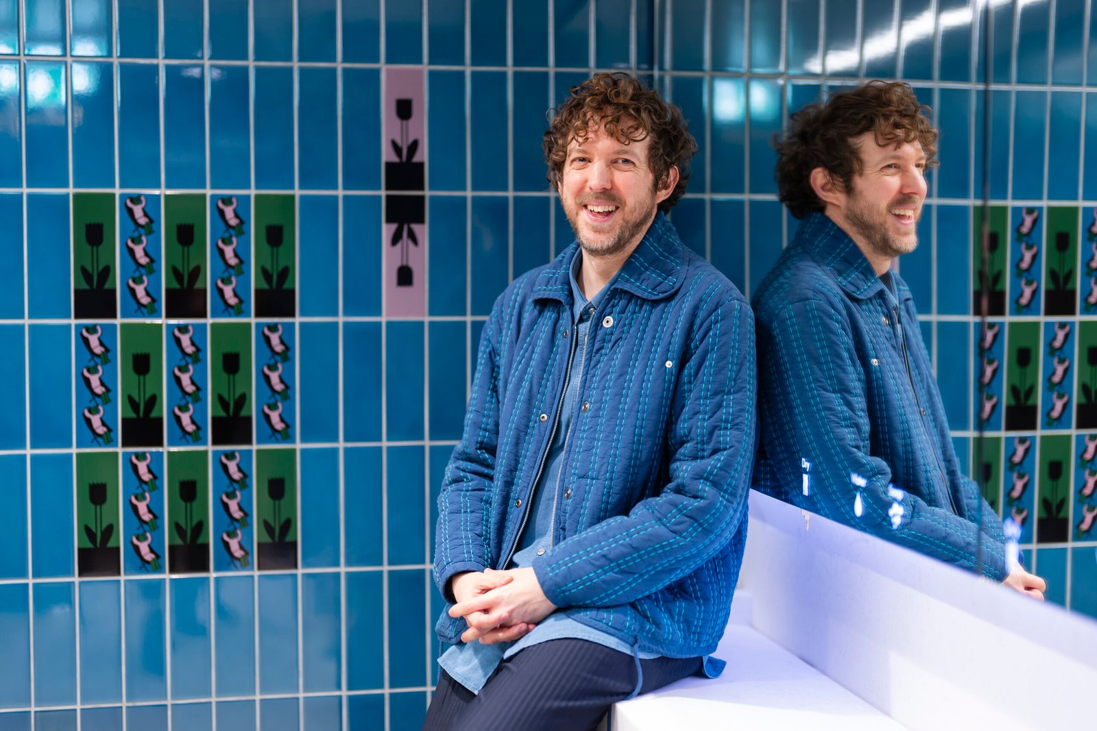 Man smiling seated against patterned tiled wall.