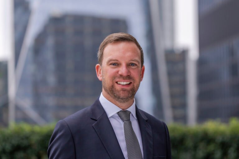 A man in a suit and tie smiles outdoors with tall, modern office buildings and greenery blurred in the background, perfectly capturing the polished style of a skilled Corporate Headshot Photographer.