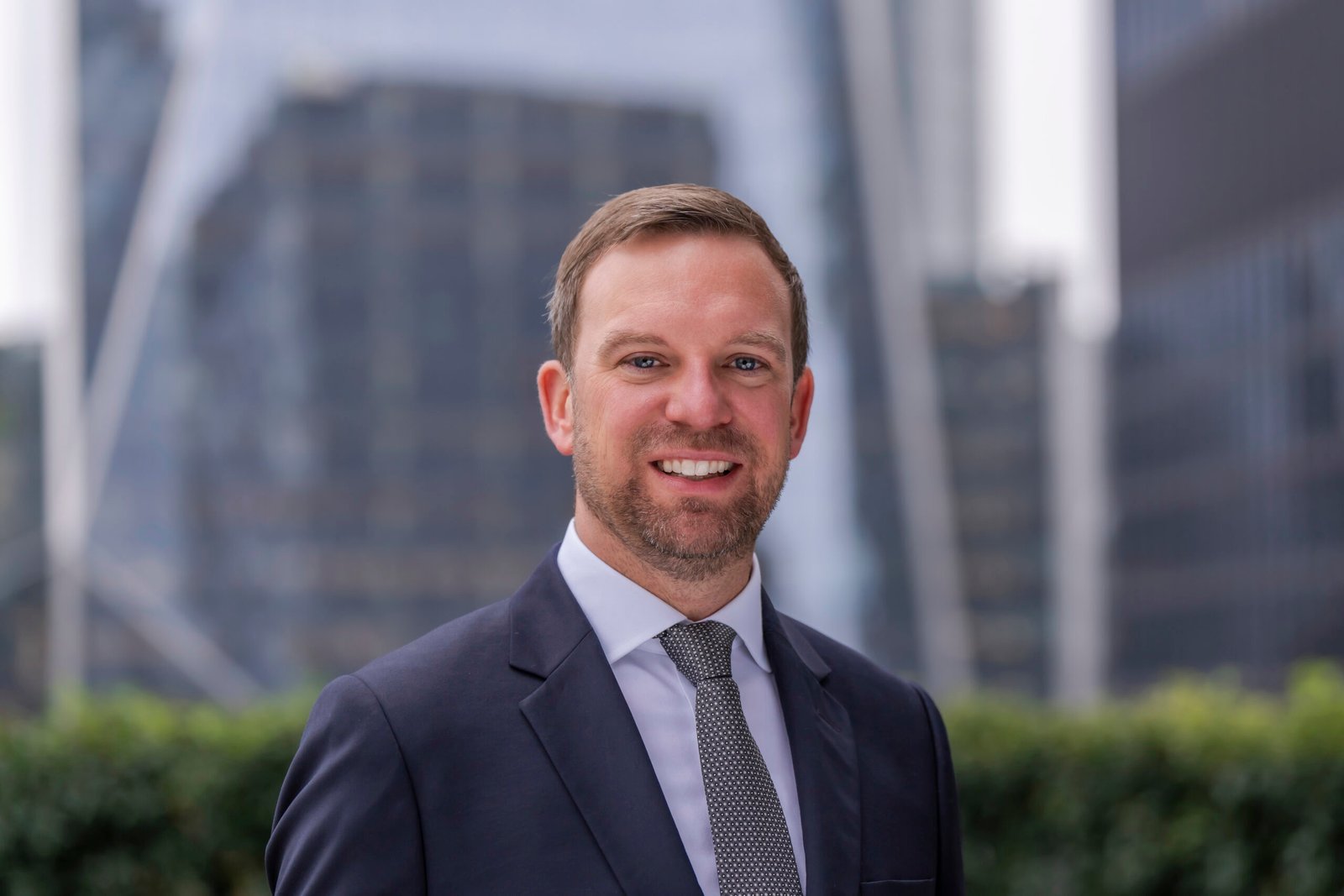 A man in a suit and tie smiles outdoors with tall, modern office buildings and greenery blurred in the background, perfectly capturing the polished style of a skilled Corporate Headshot Photographer.
