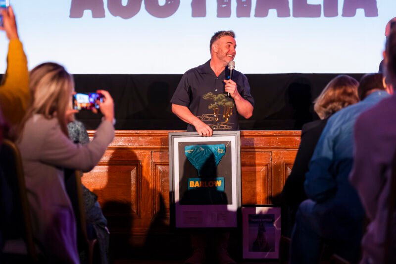 Man speaking on stage holding framed item.