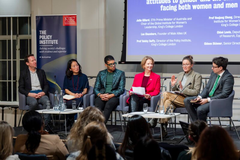 Six panellists sit on a stage in discussion at a King’s College London Policy Institute event, with an audience in the foreground and a presentation screen behind them, expertly captured by a London Conference Photographer.