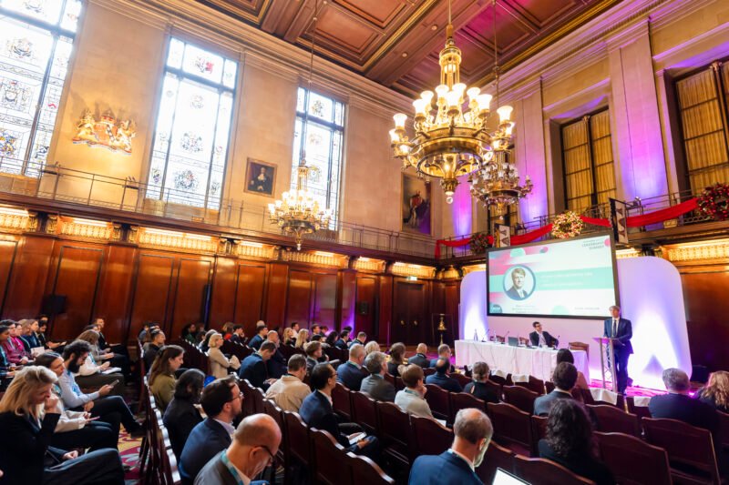 Conference in grand hall with chandeliers and stained glass.