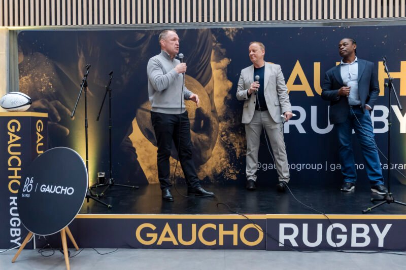 Three men speaking on stage at Gaucho Rugby event.