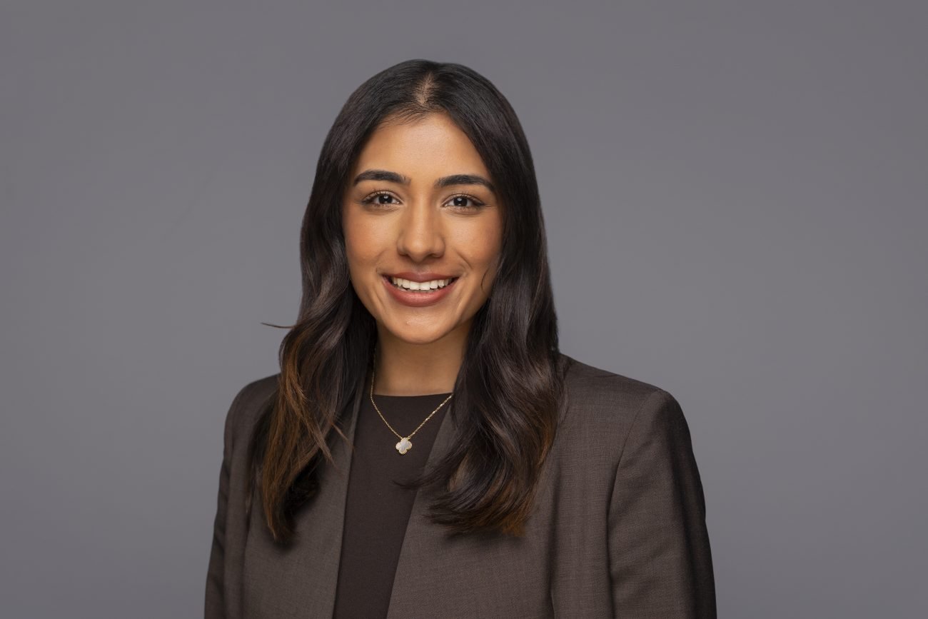A woman with long dark hair, wearing a dark blazer and a gold necklace, smiles in front of a plain grey background—an ideal example of the polished results you can expect from a professional corporate headshot photographer