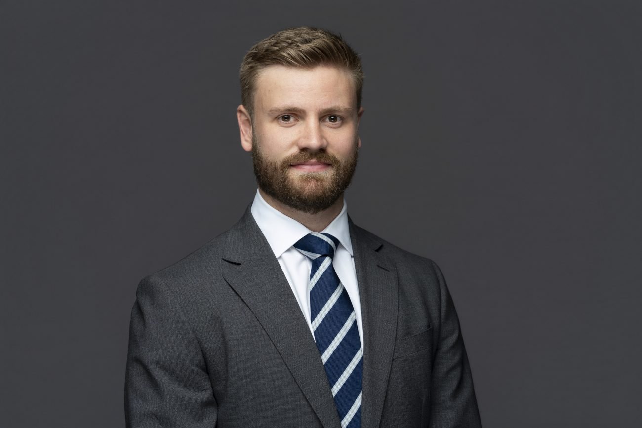 A man in a dark grey suit, white shirt, and striped tie stands against a plain dark background, looking at the camera with a neutral expression—a classic example of work by a skilled Corporate Headshot Photographer