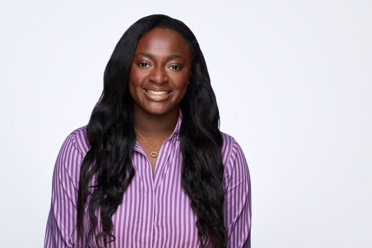 Smiling woman with long dark hair wearing a purple and white striped shirt and a gold necklace, posing in front of a plain light background, captured by a corporate headshot photographer.