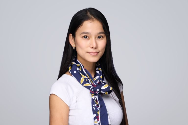 A woman with long black hair wears a white top and a blue patterned scarf, standing against a plain light grey background. Captured by a Corporate Headshot Photographer, she faces the camera with a slight smile